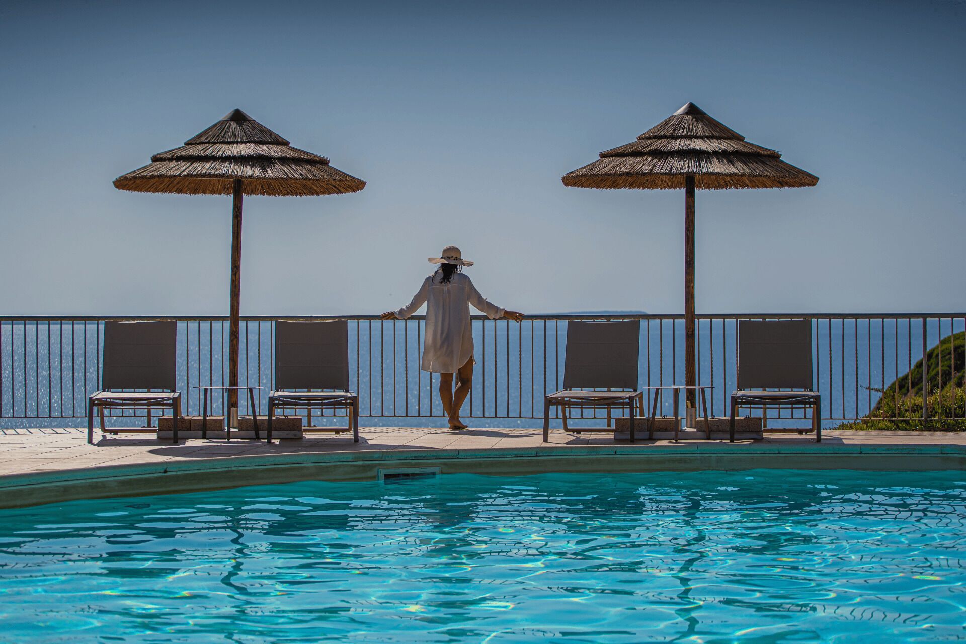 A pool area with two thatched umbrellas, lounge chairs, and a person standing by the pool.
