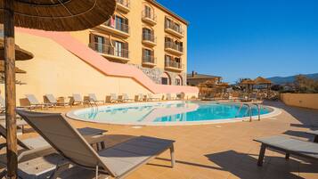 Un área junto a la piscina con sillones de descanso, una piscina y un edificio con balcones.