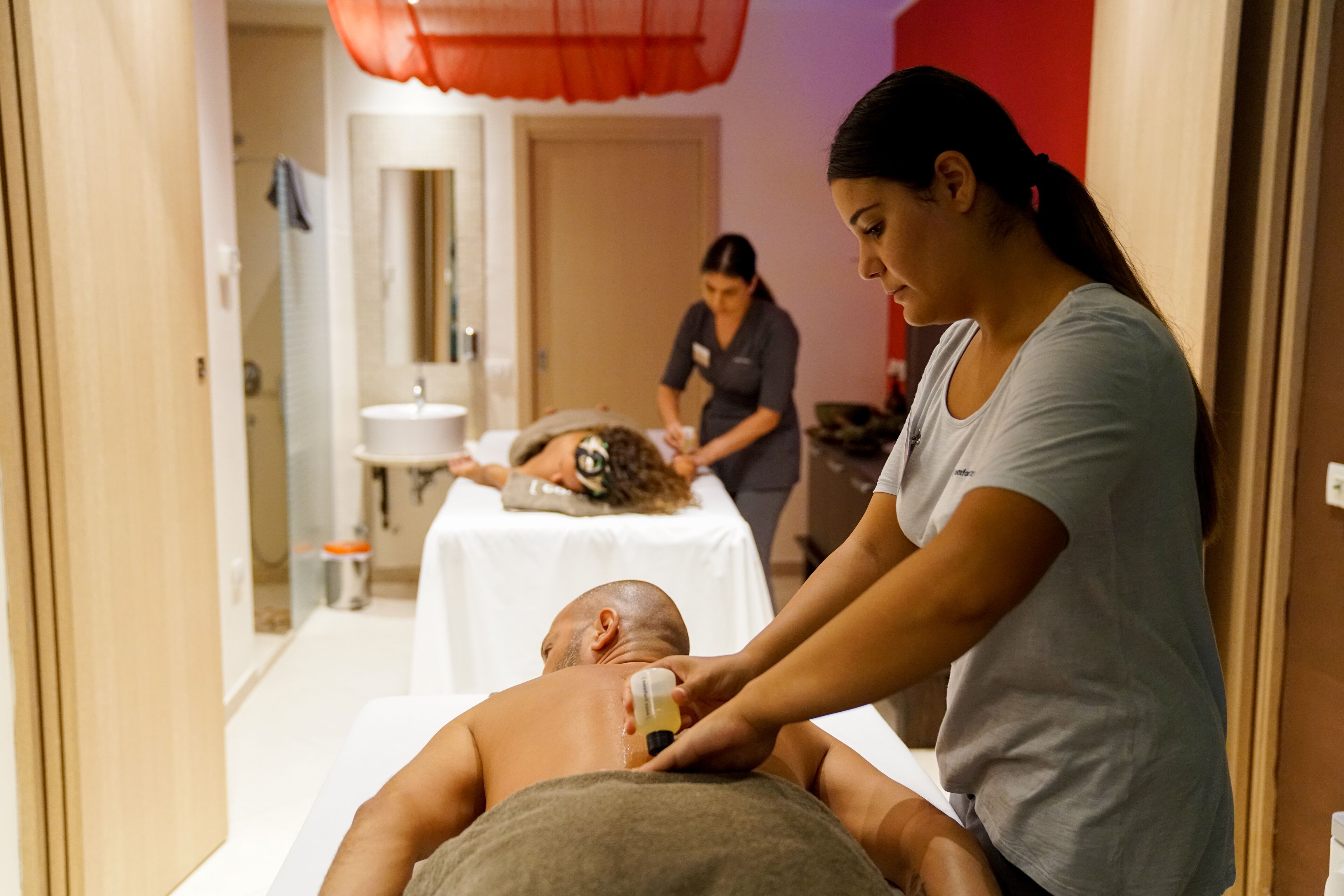 Una stanza per massaggi con un cliente sdraiato su un lettino, un terapista che esegue un trattamento e un secondo terapista sullo sfondo.
