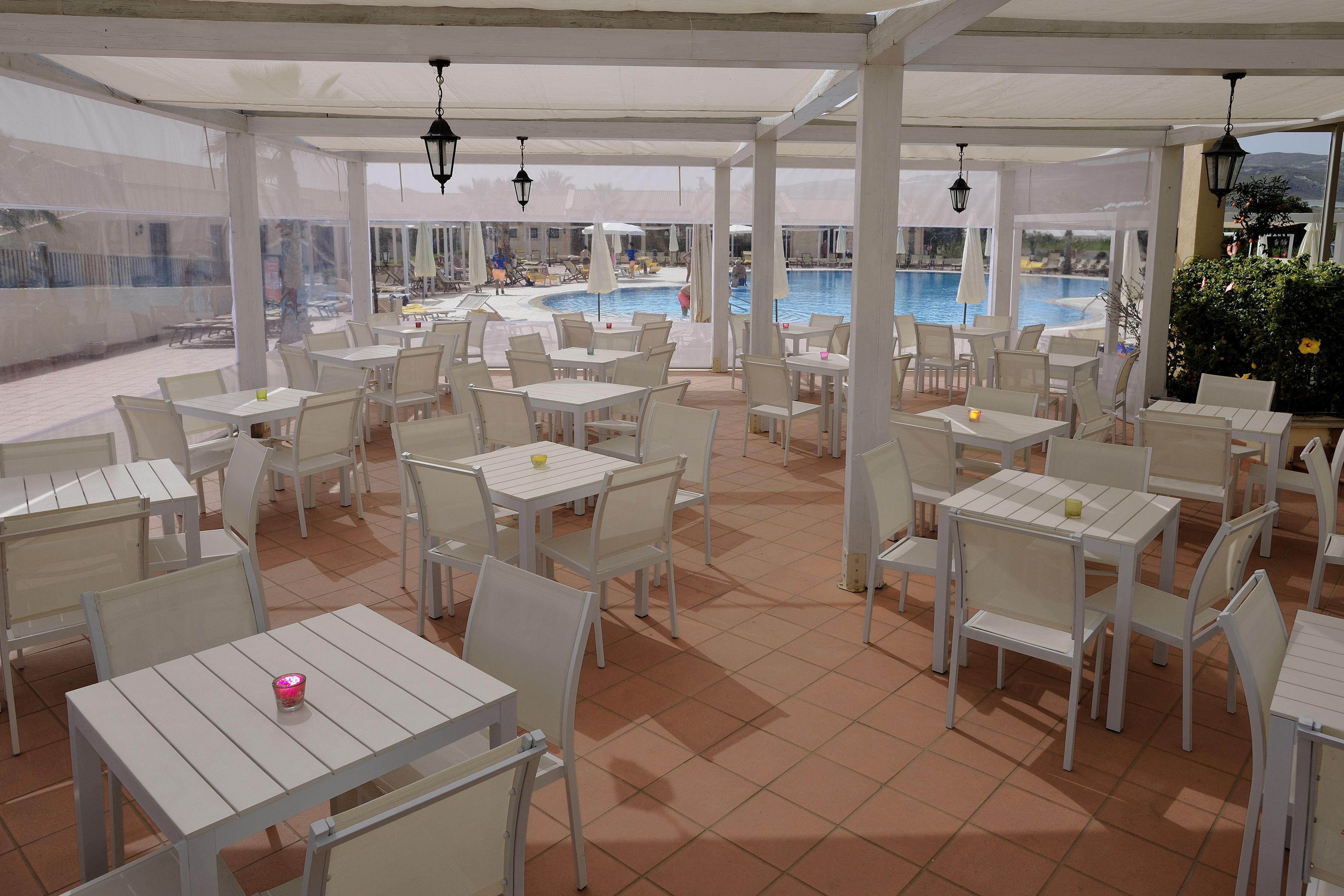 Un patio exterior con mesas y sillas blancas, una piscina al fondo y vistas del paisaje.