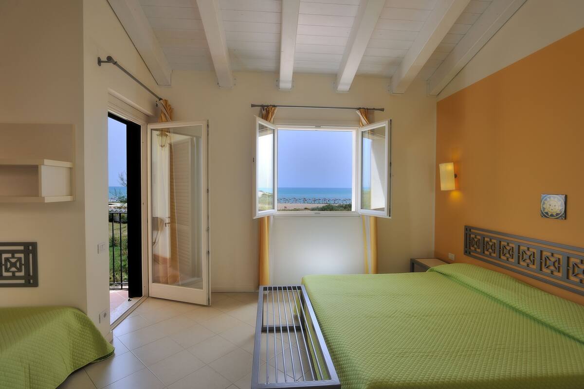 Una habitación con dos camas, una ventana que da al mar, una puerta hacia el exterior y una lámpara fijada a la pared.