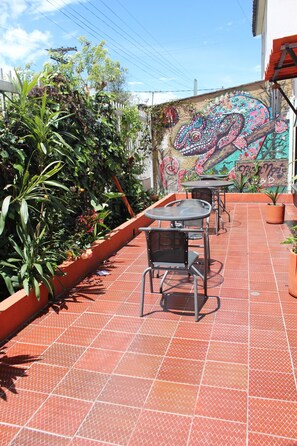 A patio with tiled flooring, metal tables, and chairs, surrounded by potted plants and a mural on the wall.