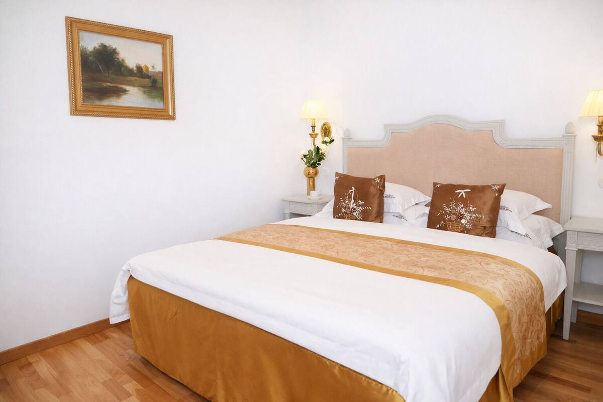 Un cuarto de hotel con una cama grande, dos mesitas de noche, una lámpara y un cuadro en la pared, además de dos lámparas de pared.