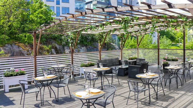 An outdoor patio with tables and chairs, a pergola with vines, and a view of buildings and greenery in the background. - Brommavik Hotel (Bromma)