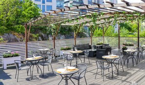 An outdoor patio with tables and chairs, a pergola with vines, and a view of buildings and greenery in the background. - Brommavik Hotel (Bromma)