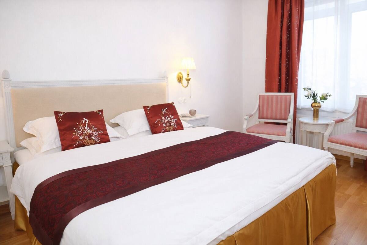 Habitación de hotel con una cama grande, una mesita de noche, una lámpara, una silla y una ventana con cortinas.