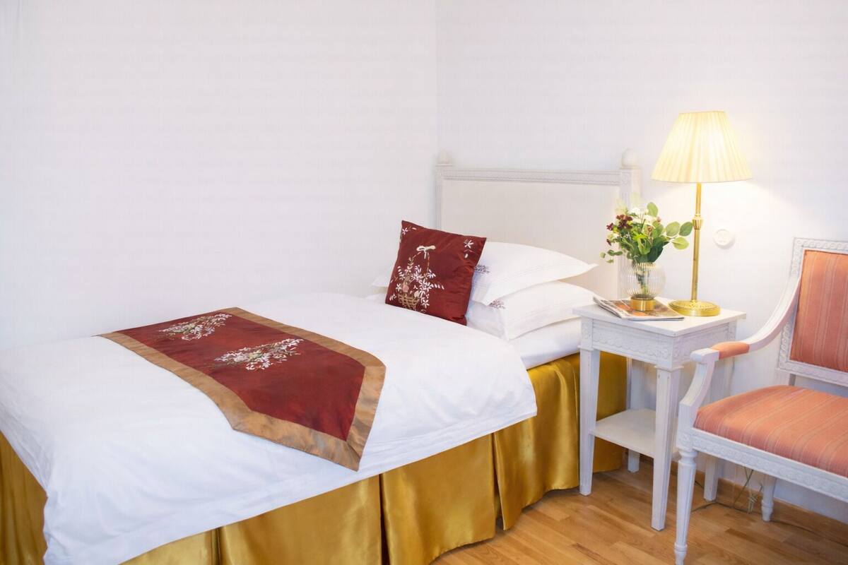 Una cama individual con cubrecolchón dorado, almohadas blancas, un plaid rojo y una mesita de noche con lámpara y flores.