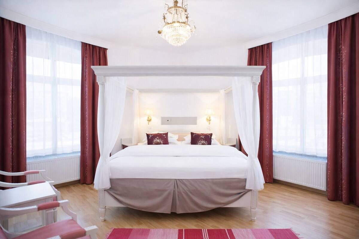 Una cama con dosel y cuatro columnas con cabecera blanca, dos lámparas de noche y un cojín rojo decorativo.