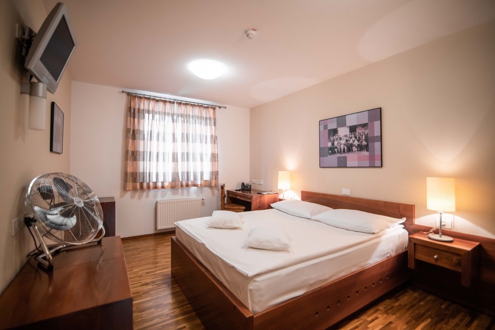 Quarto de hotel com cama, luminárias de cabeceira, ventilador, televisão e janela com cortinas.