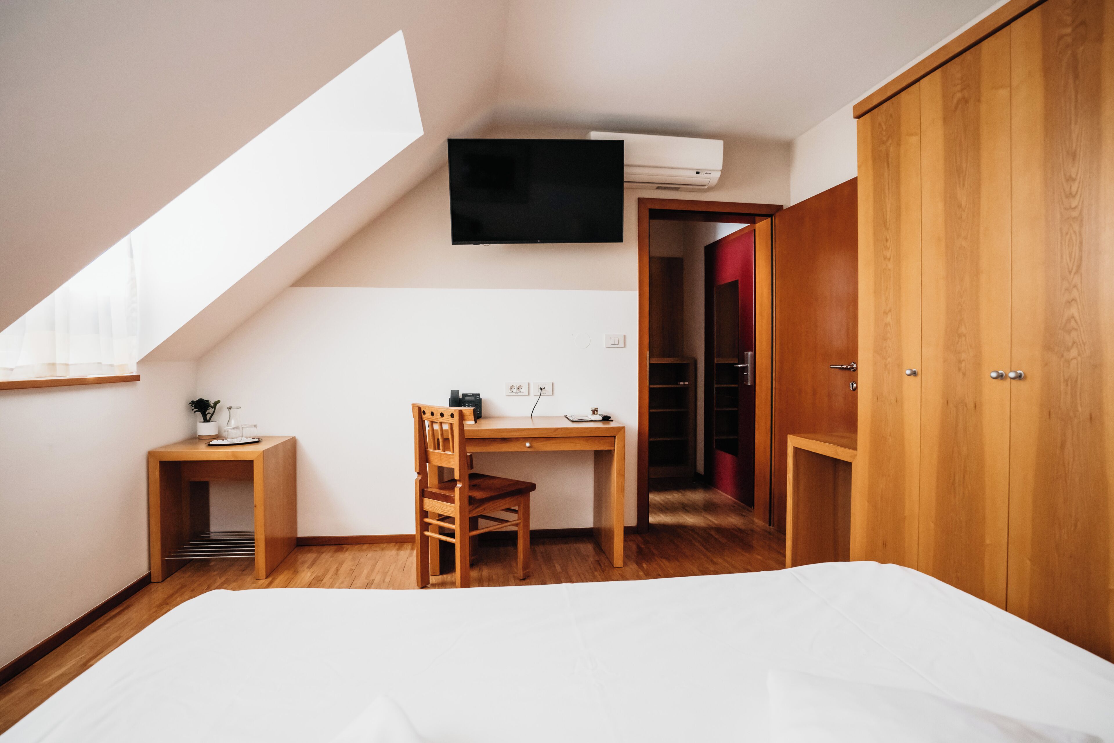 A bedroom with a sloped ceiling, a bed, a desk with a chair, a wardrobe, and a TV mounted on the wall.