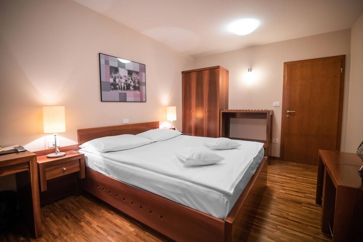 Ein ordentlich bezogenes Bett mit weißen Leinen und Kissen, ein Nachttisch aus Holz mit einer Lampe, ein Holzschrank und ein gerahmtes Foto an der Wand.