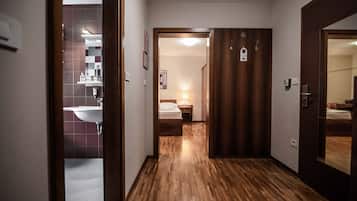 A hallway with a wooden floor leading to a bedroom with a bed and nightstand. A bathroom with a sink and mirror is visible to the left.