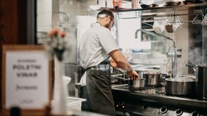 A chef in a kitchen with stainless steel appliances, pots, and a menu board visible. - Dvor Jezeršek Brnik (Cerklje na Gorenjskem)