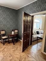 Una habitación con piso de madera en patrón de espiga, pared de color verde oscuro con diseño, una puerta de madera, una mesa redonda con sillas, y un sofá con cojines.