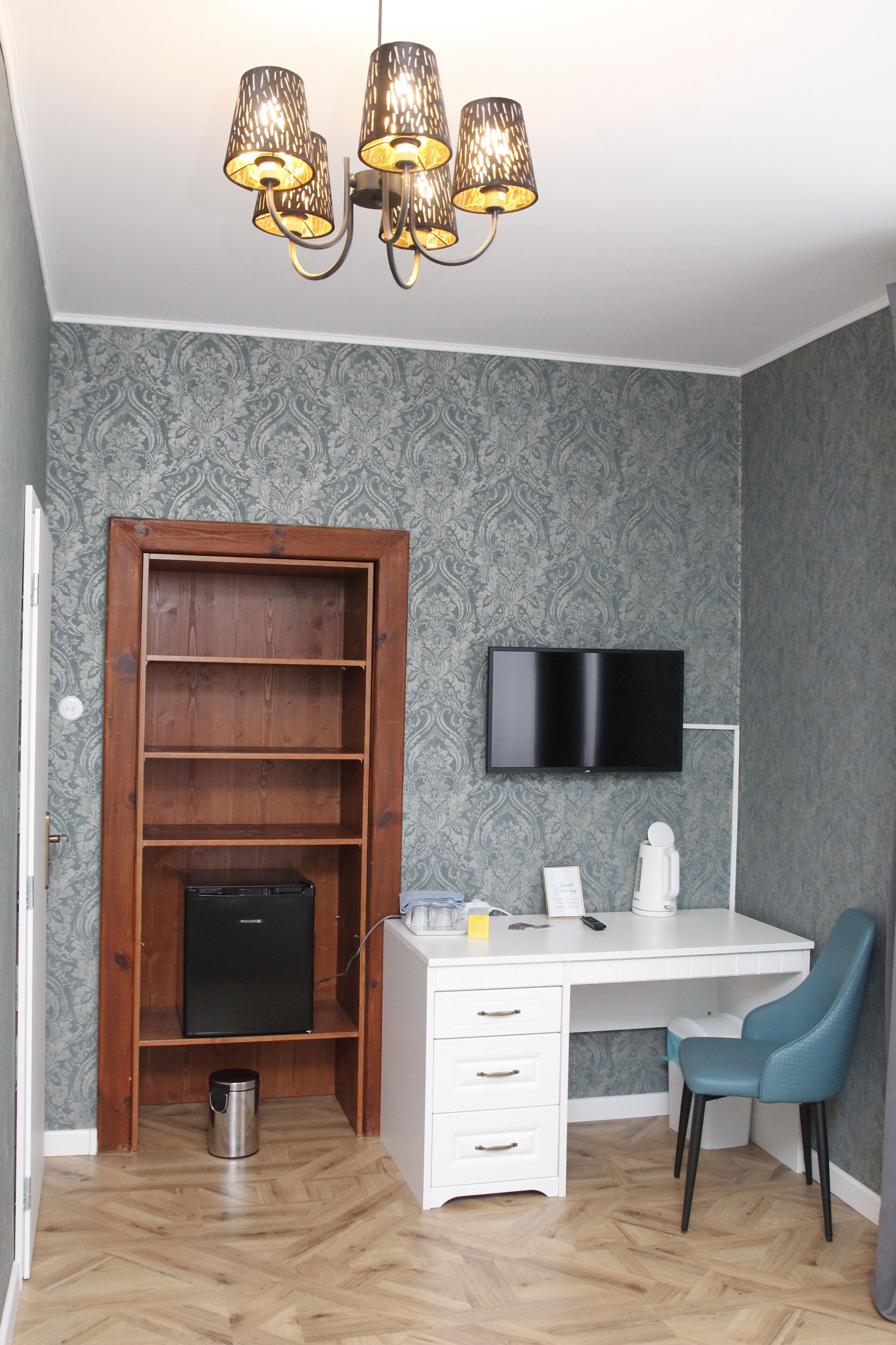 A room with a white desk, a blue chair, a television mounted on the wall, a wooden shelf unit, and a small black refrigerator.