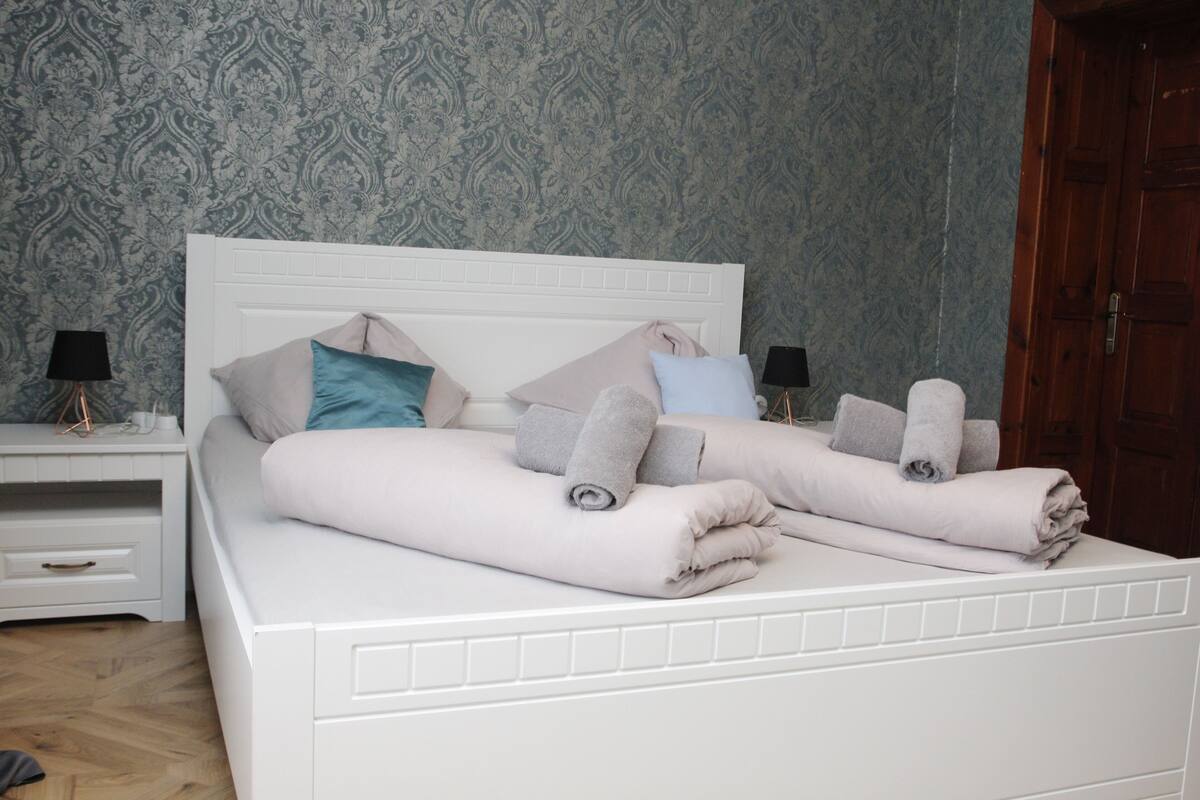 Ein ordentlich bezogenes Bett mit Kissen und Handtüchern, ein Nachttisch mit Lampe und eine Holztür in einem Zimmer mit gemustertem Wandpapier.