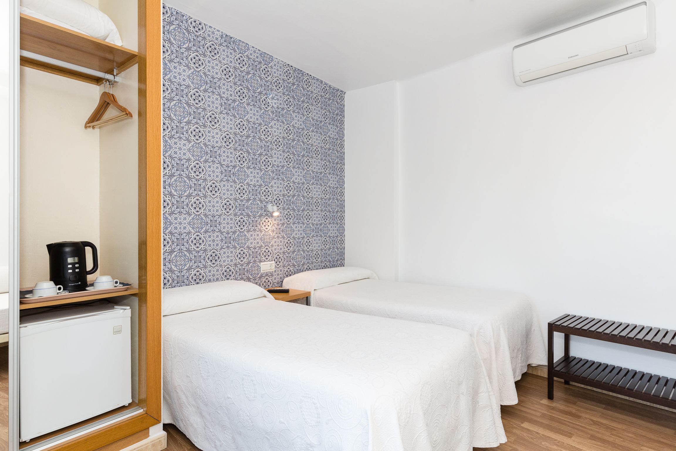 Habitación de hotel con dos camas, un armario, un refrigerador pequeño, una cafetera y un papel tapiz estampado.