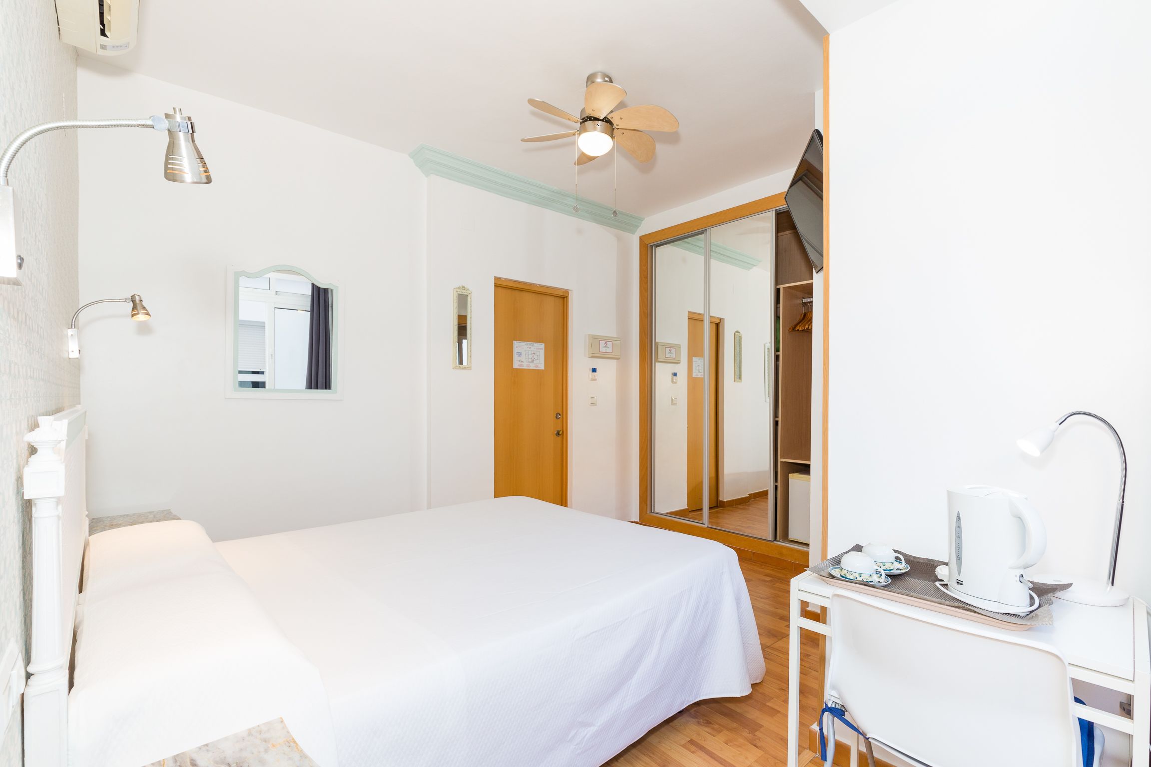 A hotel room with a bed, a desk with a kettle, a ceiling fan, and a mirror.