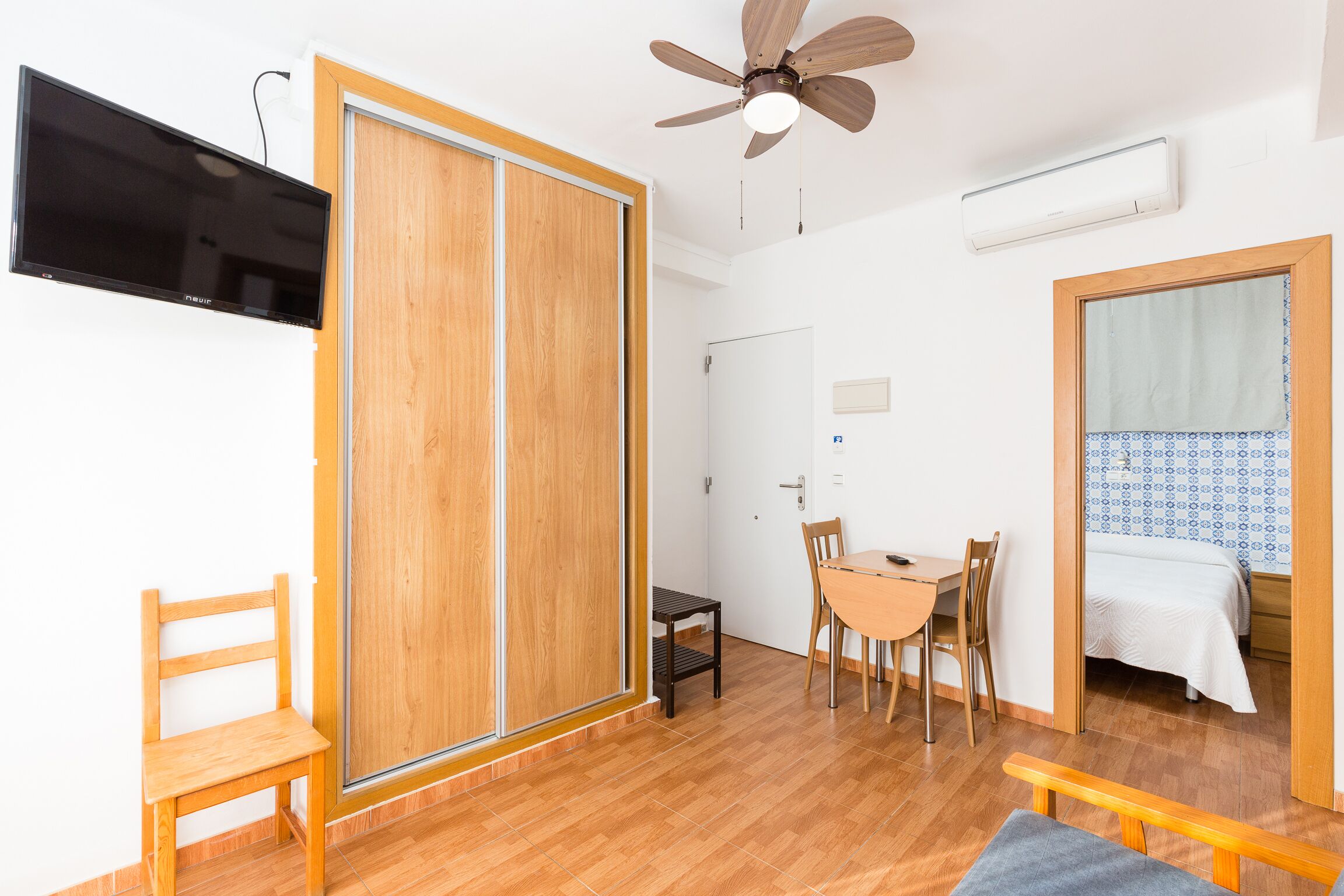 Una habitación con piso de madera, ventilador de techo, televisor montado en la pared, cama con cubrecama estampado y una zona de comedor con mesa y sillas.