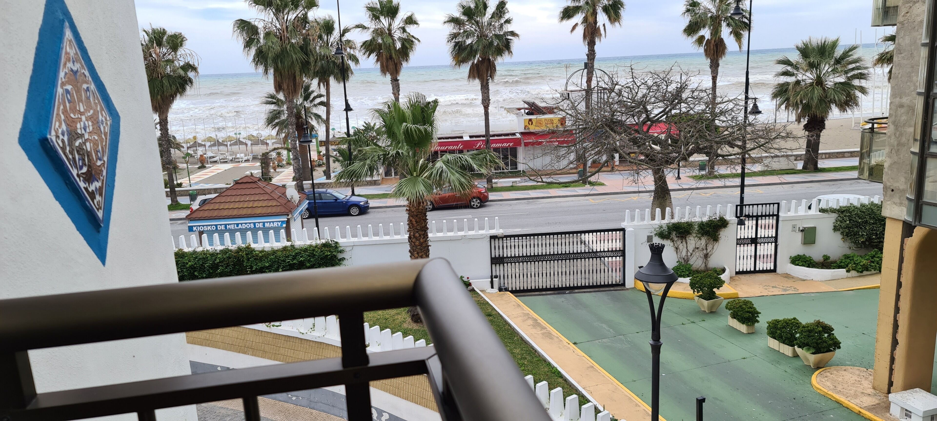 A balcony with a view of the beach, palm trees, and a street with cars and a restaurant.