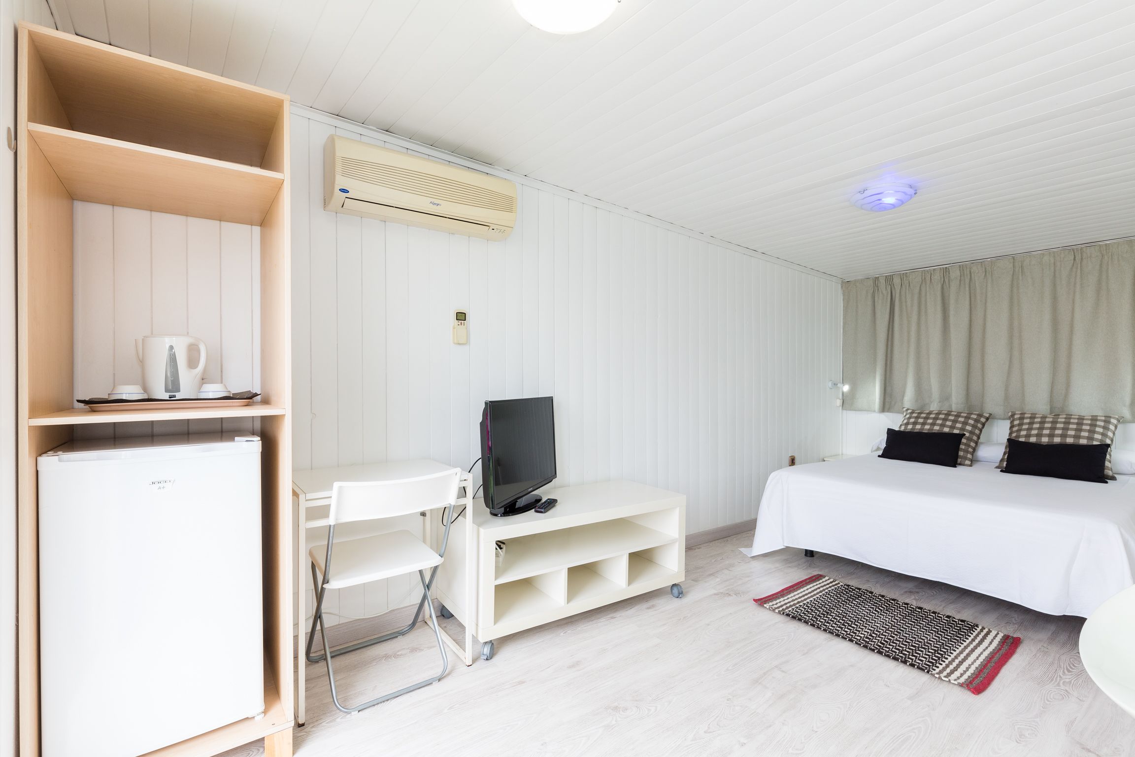 A hotel room with a bed, a TV on a stand, a desk with a chair, a small fridge, and a window with curtains.