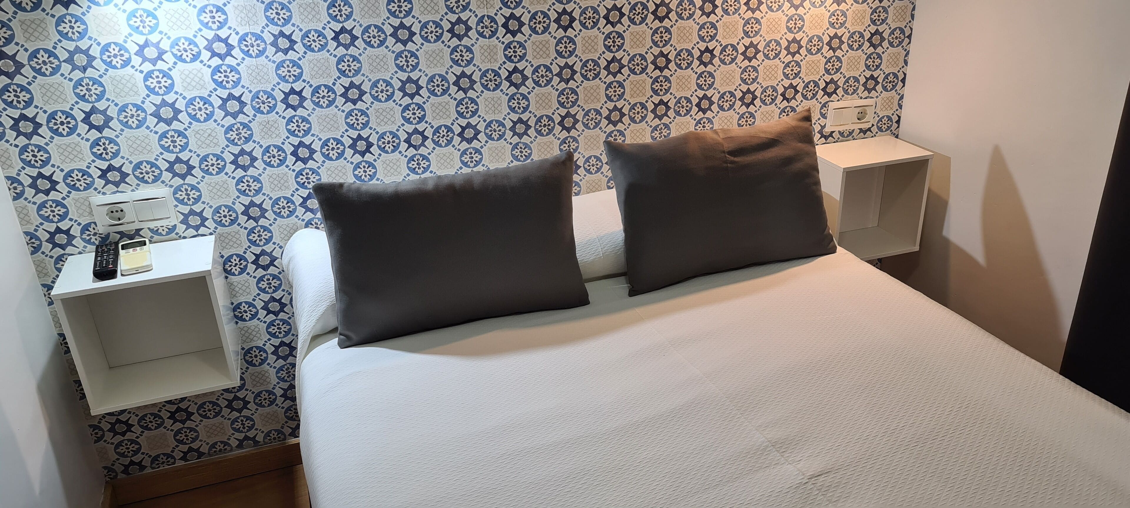 A neatly made bed with two pillows, a bedside table with a lamp, and a wall with a blue and white patterned wallpaper.