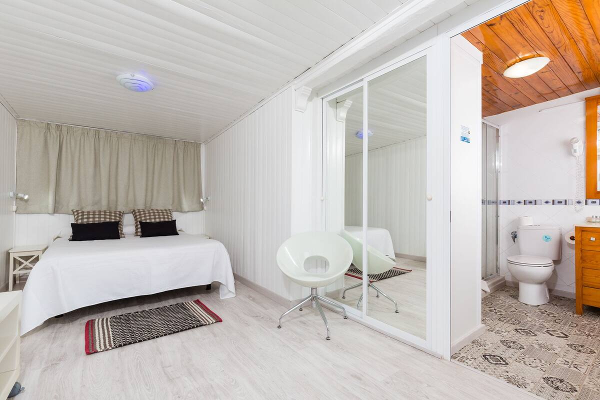 Un dormitorio con cama, una silla blanca, un baño con inodoro y lavamanos, y un suelo con diseño.