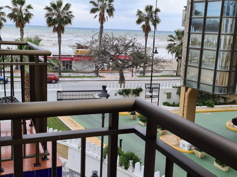 Un balcón con vista a la playa y palmeras, un coche rojo aparcado en la calle, y un edificio con una ventana y una puerta.