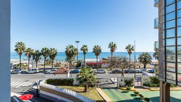 A view from a balcony overlooking a beach with palm trees, a road with cars, and a building with a balcony.