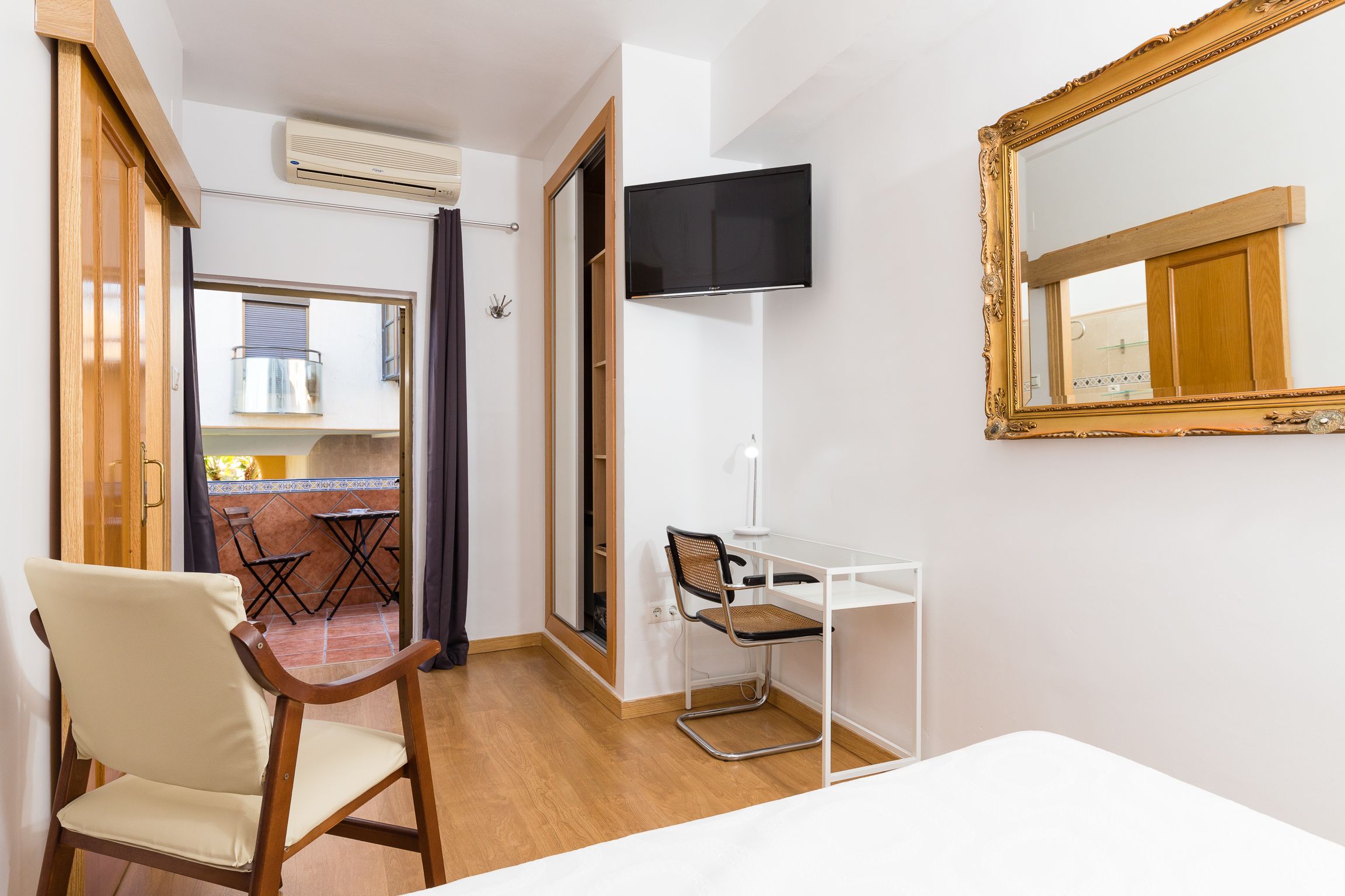 A hotel room with a wooden chair, a desk, a television mounted on the wall, and a large mirror.
