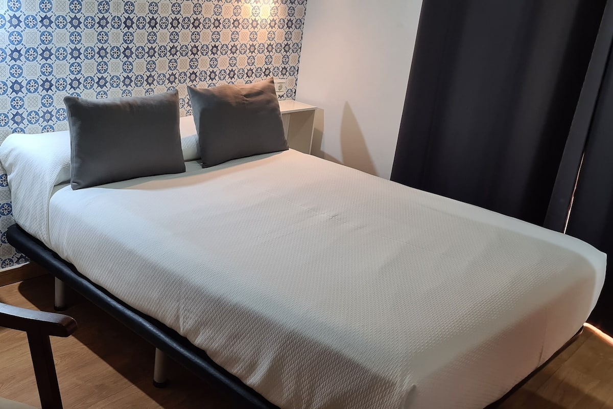 Una cama bien hecha con una cubierta blanca y dos almohadas, una silla de madera y una pared con un diseño llamativo al fondo.