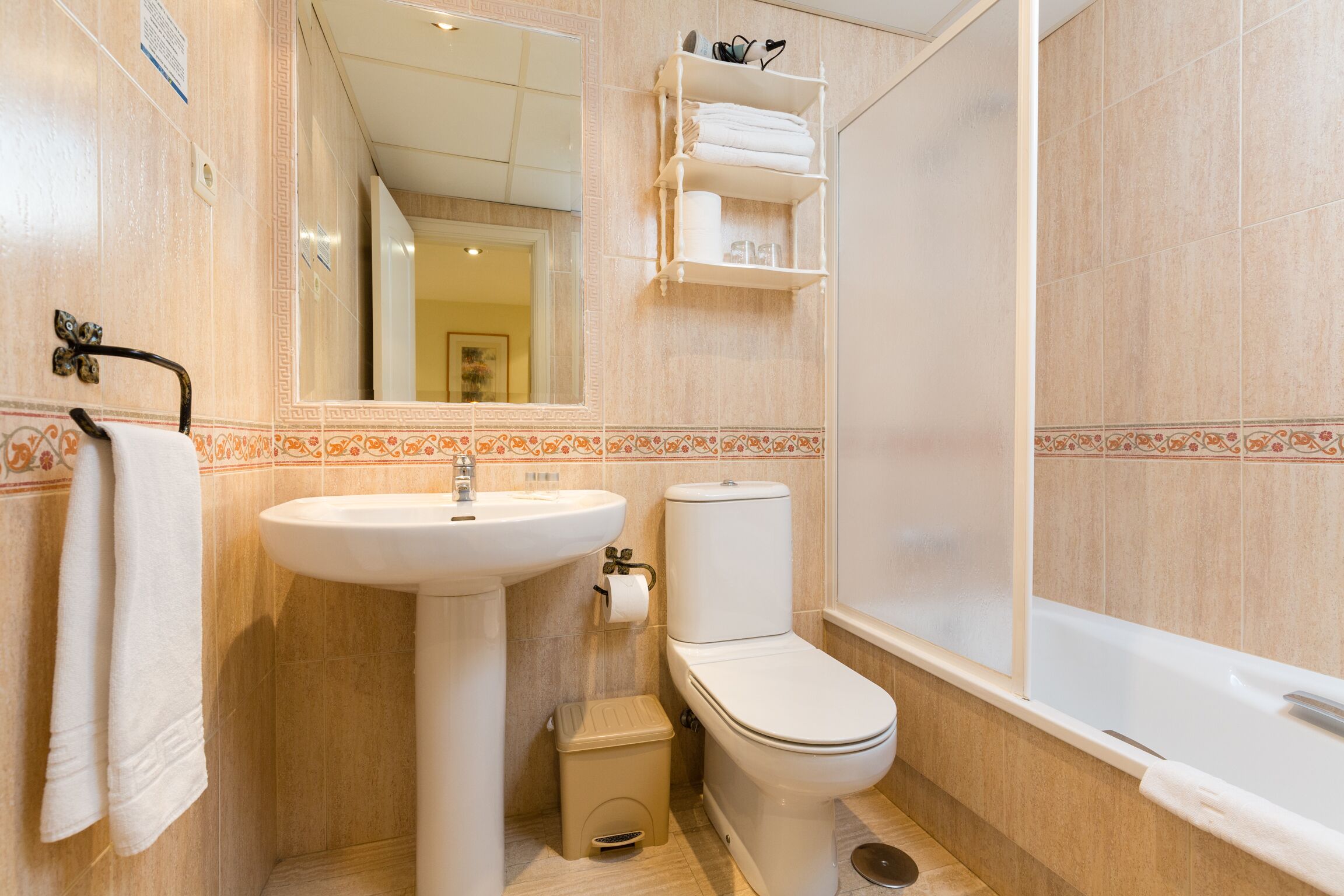 Un baño con lavamanos, inodoro y bañera. Hay un perchero y una repisa con artículos de aseo.