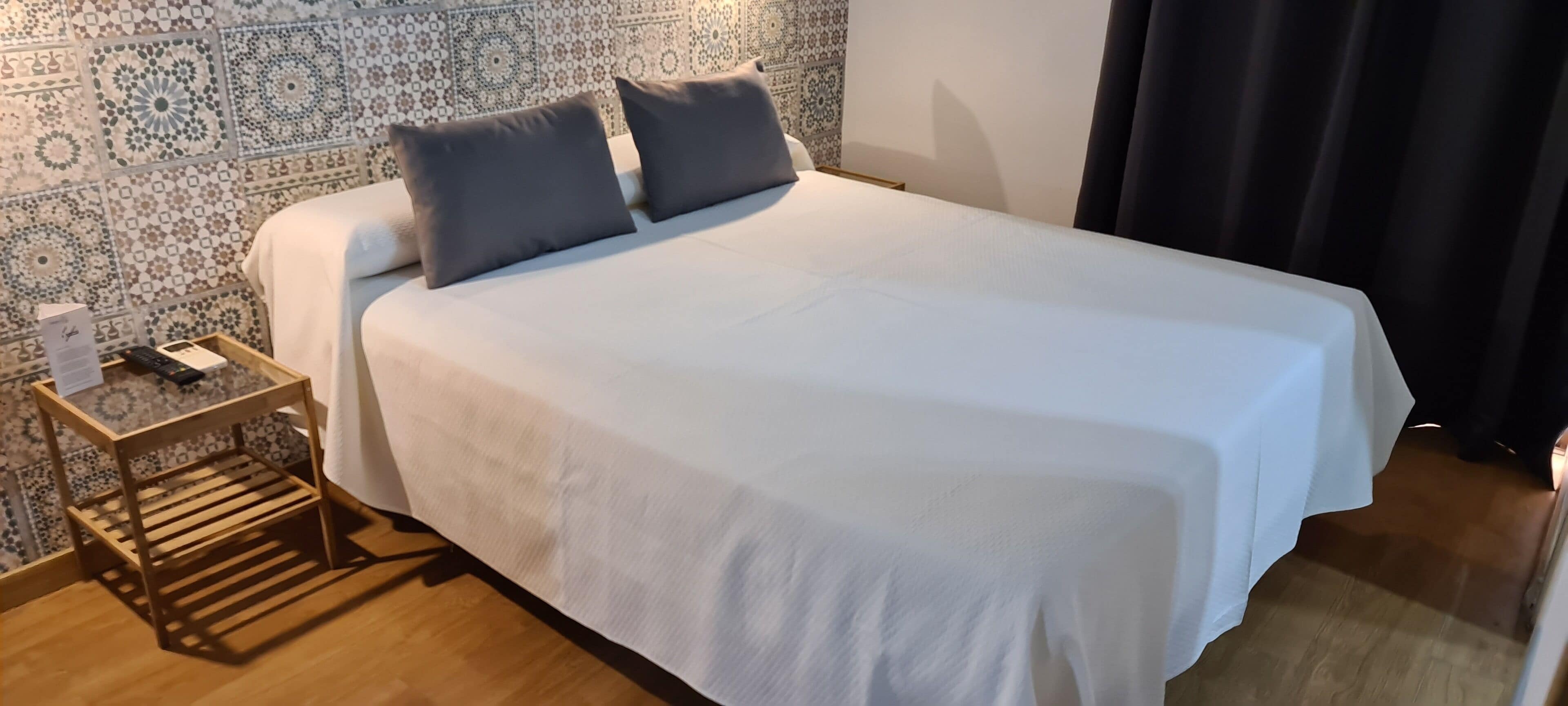 A neatly made bed with a white duvet and two gray pillows, a wooden nightstand with a lamp, and a patterned wall behind the bed.