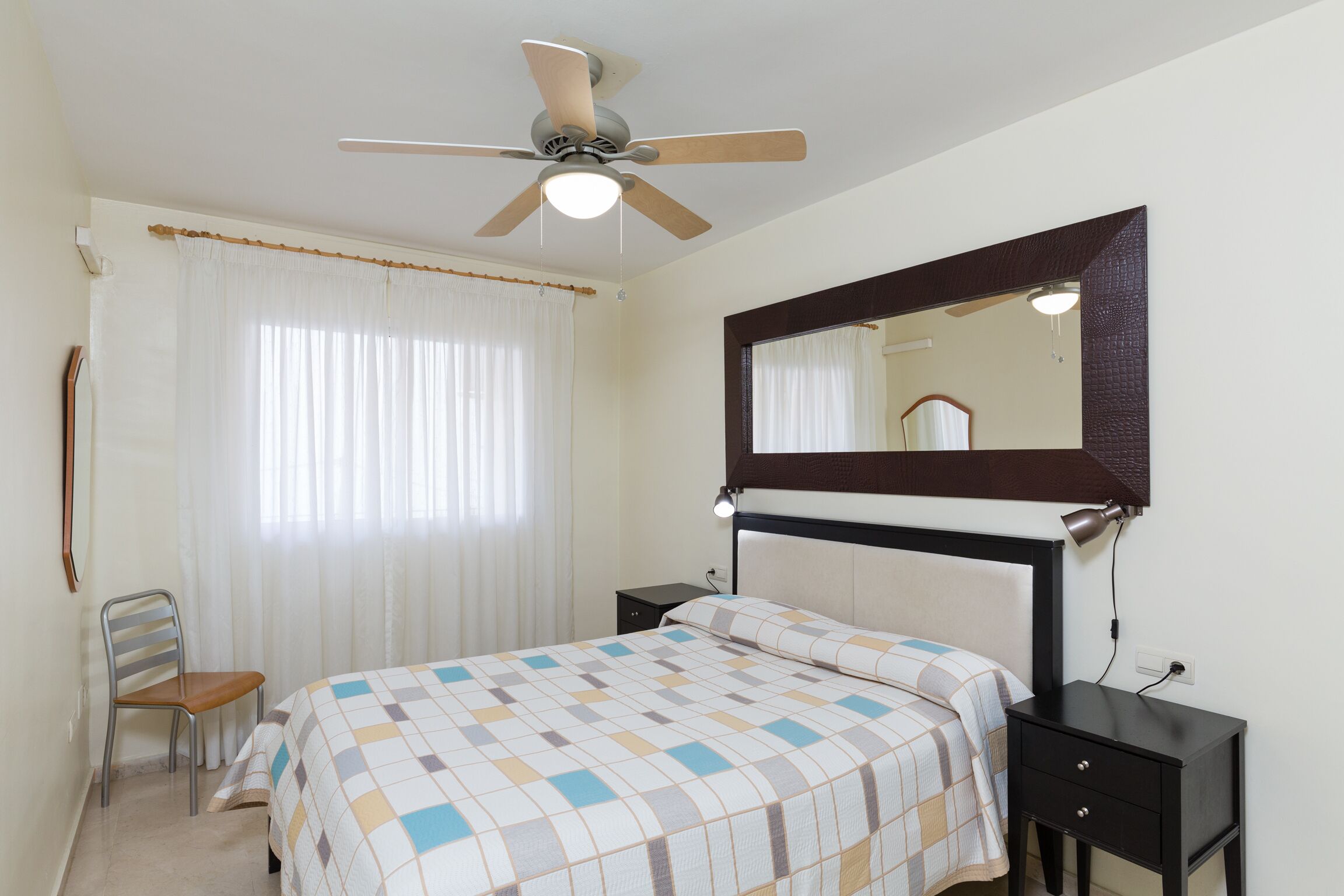 A bedroom with a bed, a chair, a nightstand, a mirror, and a ceiling fan.