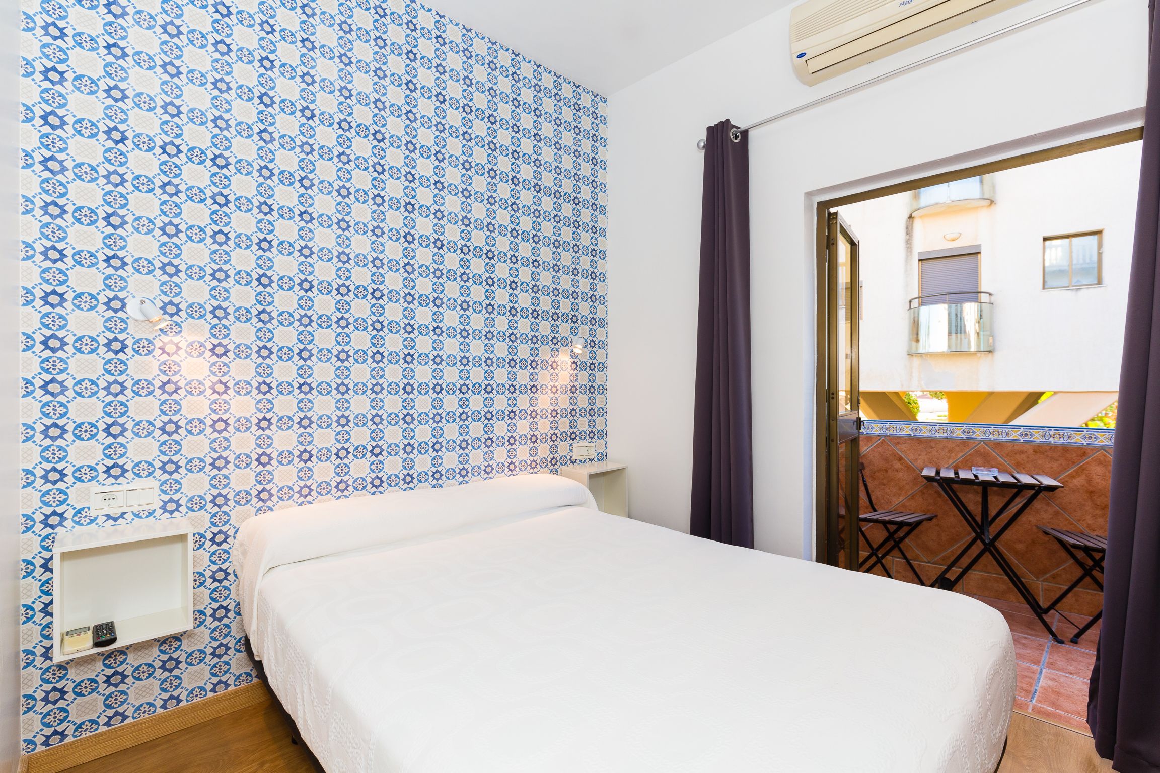 A room with a bed, a small table, and a chair, a patterned wall, and an open door leading to an outdoor area with a table and chairs.