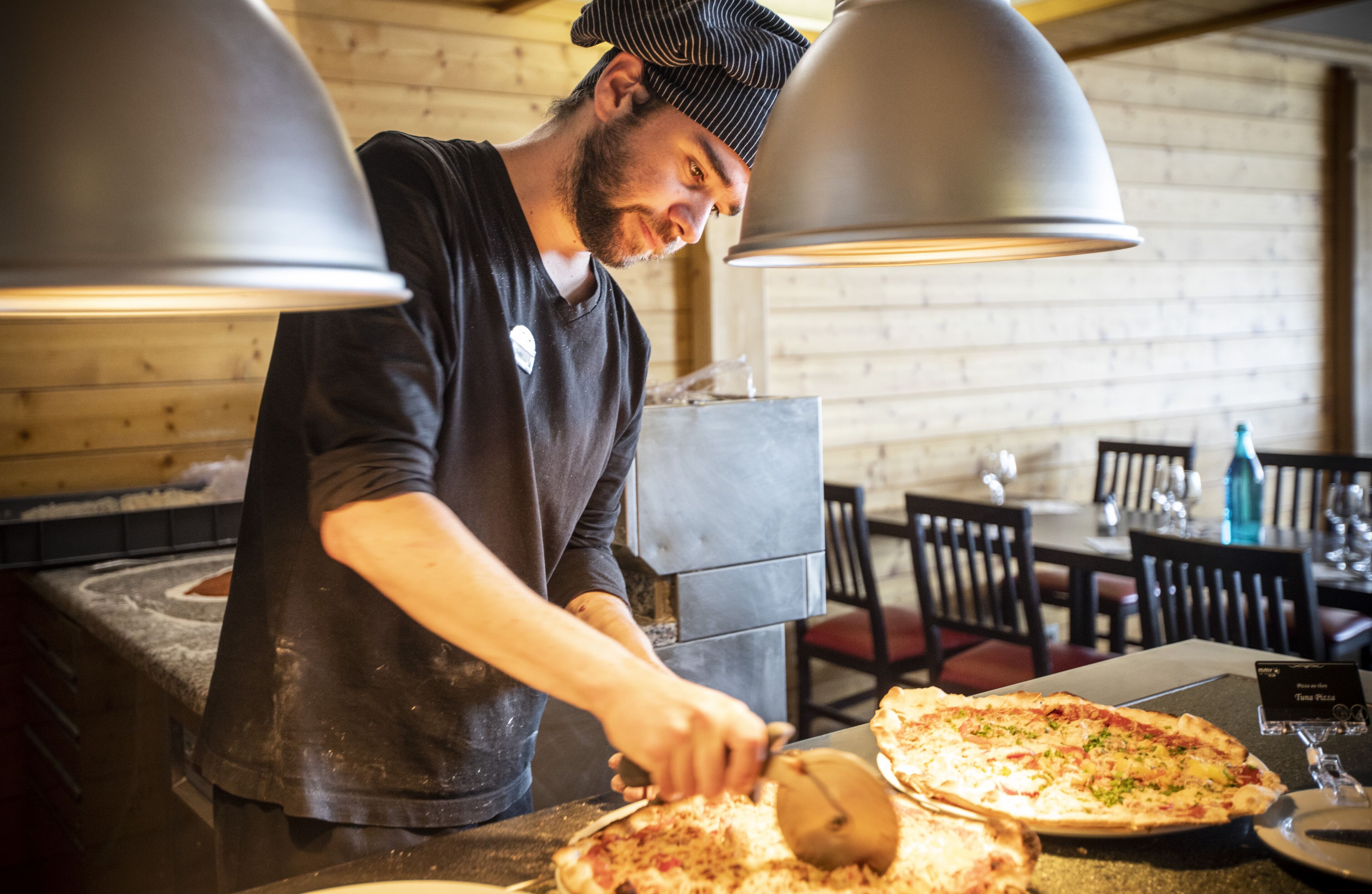 A chef slicing pizza in a rustic kitchen with wooden walls and pendant lights.