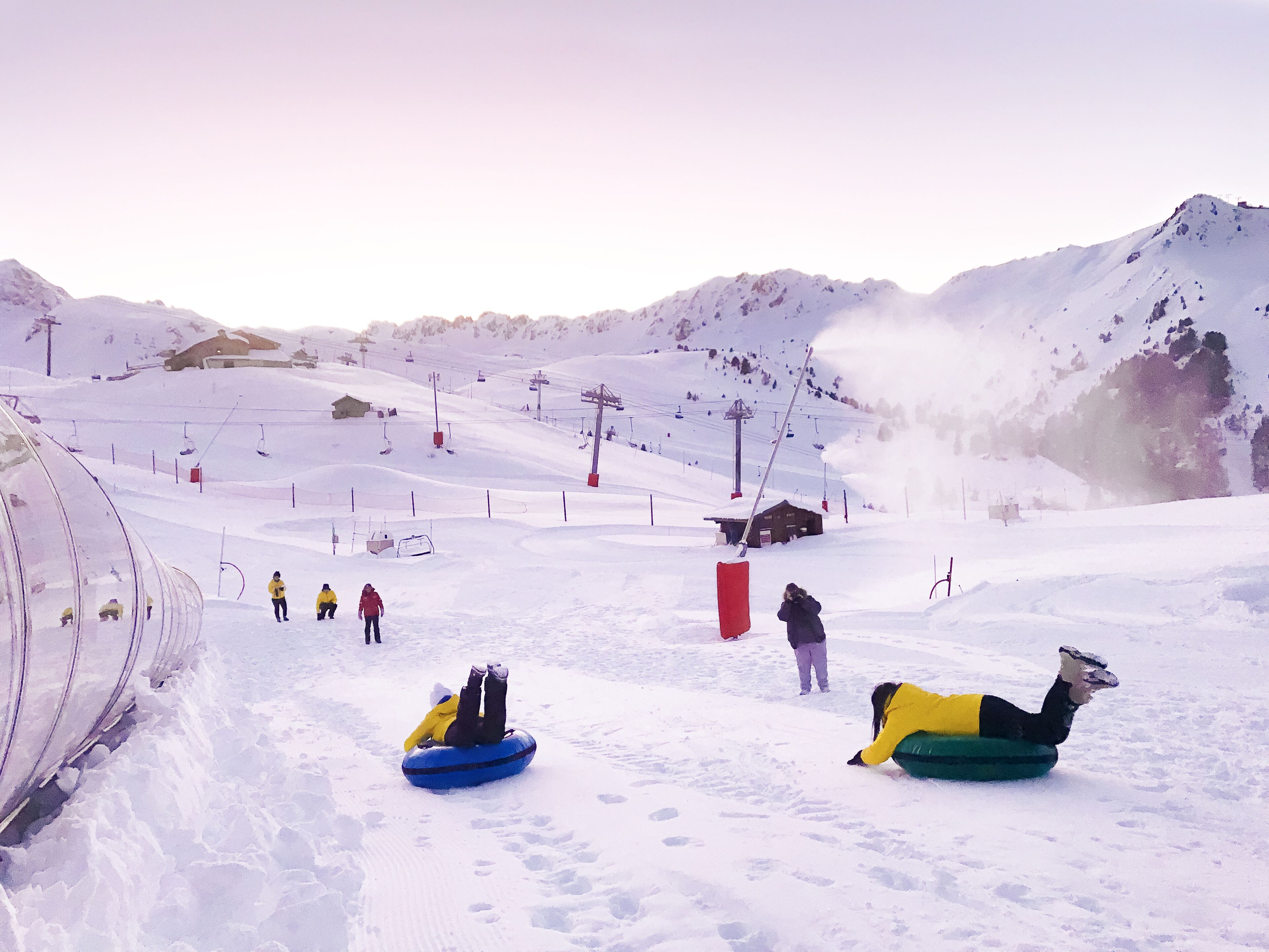 A snowy ski slope with a ski lift, a wooden cabin, and people on snow tubes.