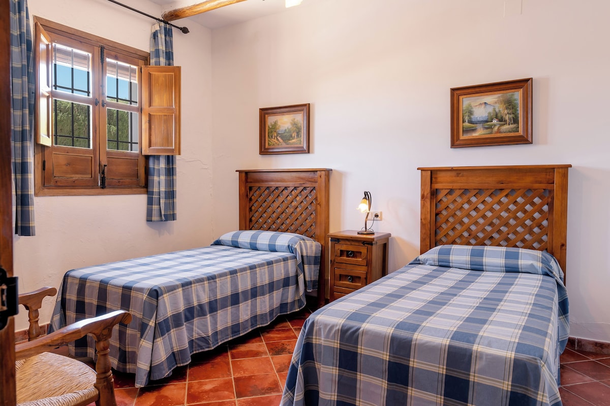 A room with two beds, a wooden headboard, a small bedside table, a chair, and a window with curtains.