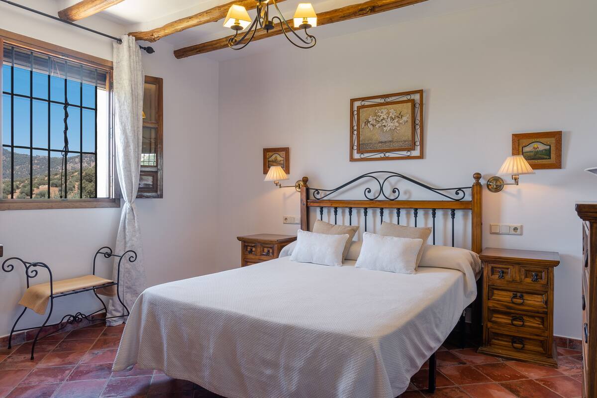 A bedroom with a large bed, wooden headboard, two bedside tables, a bench, and a window with curtains.