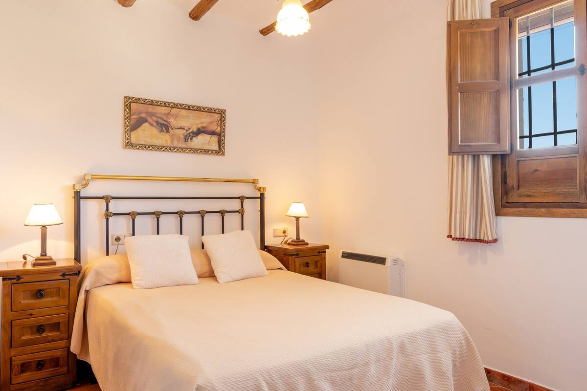 A neatly made bed with a headboard, two bedside tables with lamps, a radiator, a window with curtains, and a framed picture on the wall.