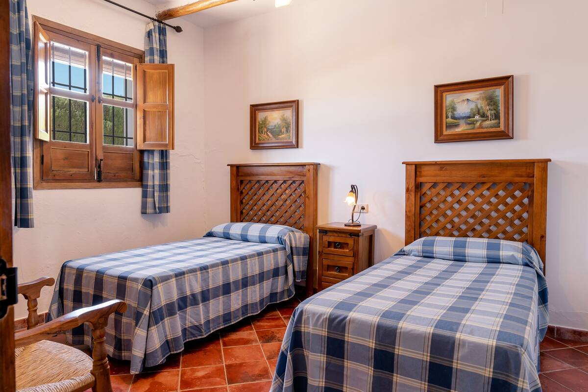 A room with two beds, a wooden headboard, a nightstand, a chair, a window with curtains, and two wall-mounted paintings.