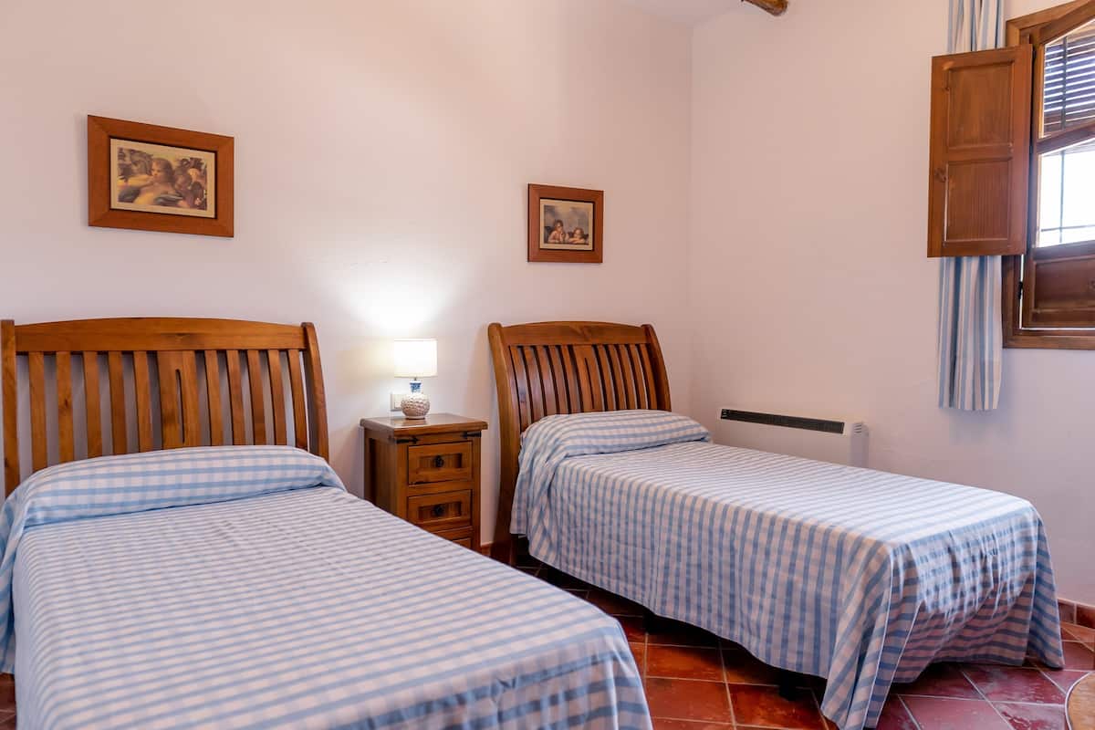 A bedroom with two single beds, a nightstand with a lamp, and a window with blinds.