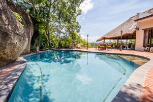 Uma piscina com uma parede de pedra, rodeada por vegetação exuberante e uma construção com telhado de palha.