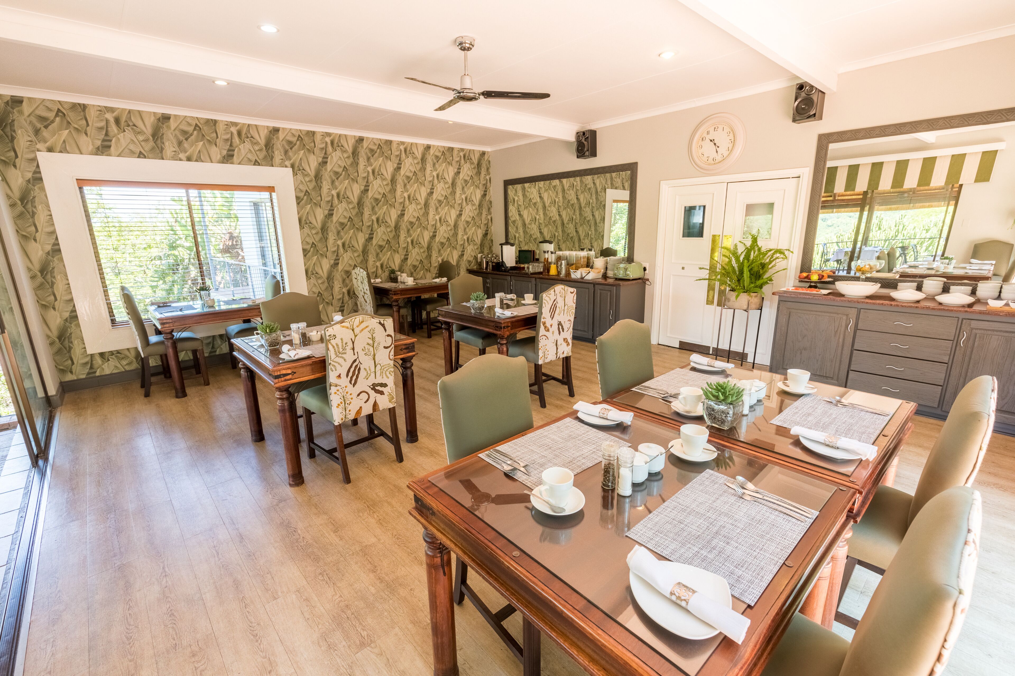 Un coin repas avec une table en bois dressée pour quatre personnes, entourée de chaises, d’un ventilateur de plafond et d’une fenêtre équipée de stores.