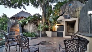 An outdoor patio area with stone flooring, metal chairs, a stone staircase, and a thatched-roof structure. - La Roca Guest House (Mbombela)