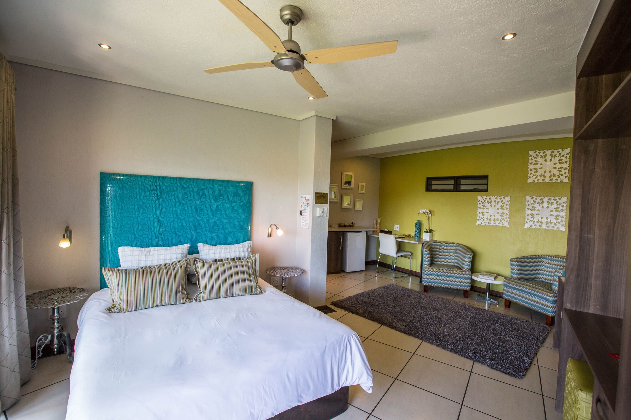 A hotel room with a bed, a ceiling fan, a kitchenette, and two chairs.