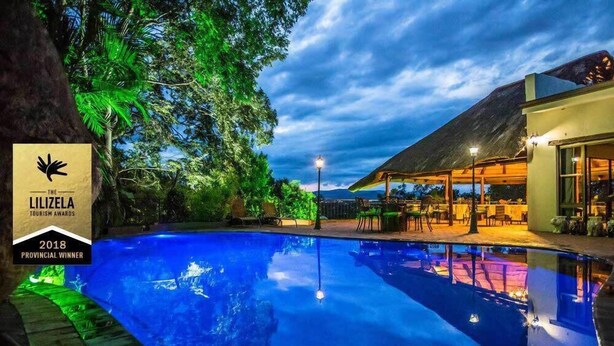 A swimming pool with a thatched-roof structure and outdoor seating, surrounded by lush greenery. - La Roca Guest House (Mbombela)