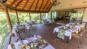 An outdoor dining area with tables set for breakfast, a thatched roof, and a view of the surrounding greenery.