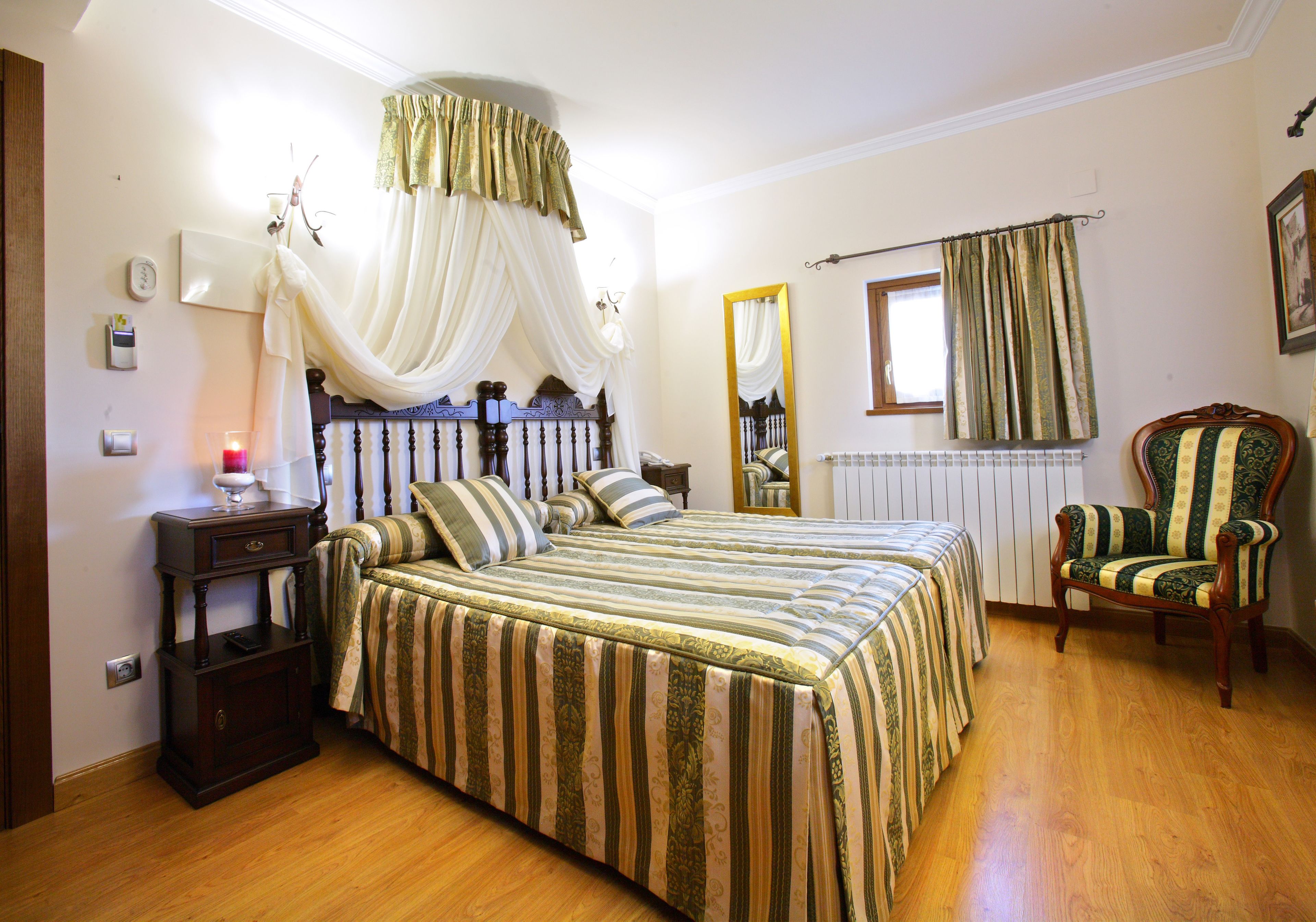 A bedroom with a large bed, a nightstand, a chair, a mirror, and a window with curtains.