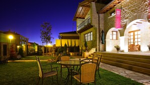 A courtyard with a dining area featuring wicker chairs and a table, surrounded by stone walls and a well-lit building with a balcony. - El Juglar (Olite)